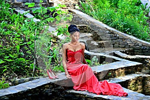 African American woman sitting in the Park posing against the backdrop of green plants on the rocks in a red dress with dreadlocks