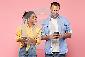 African american woman spying on her smiling boyfriend who using cellphone, standing together over pink background