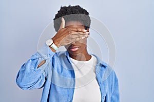 African american woman standing over blue background covering eyes with hand, looking serious and sad