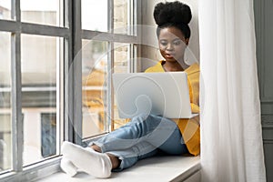 African American woman student sitting on windowsill, working doing remote job on laptop, learning using online course. Self-