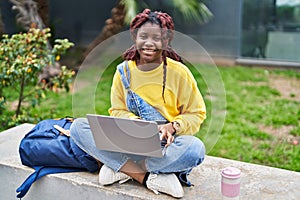 African american woman student using laptop sitting on bench at campus park