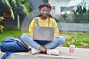 African american woman student using laptop sitting on bench at campus park