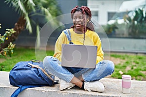 African american woman student using laptop sitting on bench at campus park