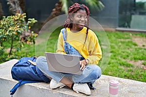 African american woman student using laptop sitting on bench at campus park