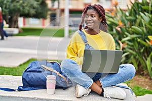 African american woman student using laptop sitting on bench at park