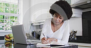 African american woman taking notes and using laptop in the kitchen while working from home