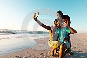 African american woman taking selfie with husband and son over smartphone at beach against sky