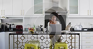 African american woman talking on smartphone and using laptop in the kitchen while working from home