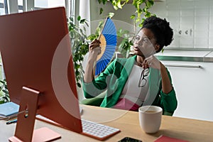 African businesswoman uses hand fan sits at desk with computer in summer day, suffers from heat.