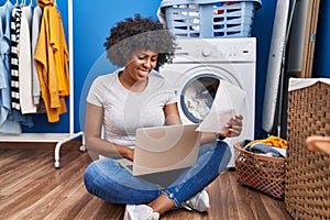 African american woman using laptop reading paperwork waiting for washing machine at laundry room