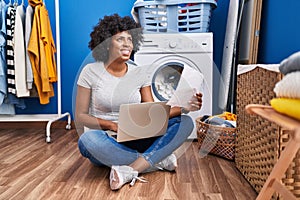 African american woman using laptop reading paperwork waiting for washing machine at laundry room