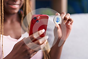 African american woman using smartphone holding bitcoin at home