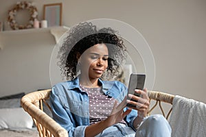 African American woman using smartphone, relaxing in cozy armchair