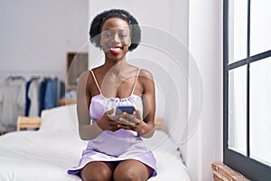 African american woman using smartphone sitting on bed at bedroom