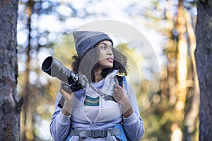 African American woman walking in the forest with a camera looking by side.