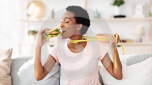 African American woman wanting to eat cheeseburger, pulling herself away by tape measure at home, panorama