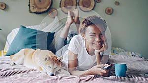 African American woman is watching TV holding remote control pressing buttons and laughing while her pet dog is lying