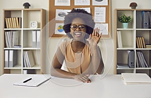 African american woman waving hand looking at camera sitting at office desk
