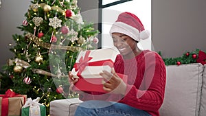 African american woman wearing christmas hat unpacking gift with surprised face at home