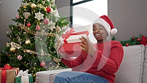 African american woman wearing christmas hat unpacking gift with surprised face at home