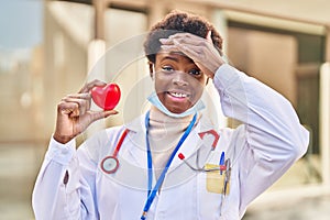 African american woman wearing doctor uniform holding heart stressed and frustrated with hand on head, surprised and angry face