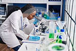 African american woman wearing scientist uniform writing on clipboard using microscope laboratory