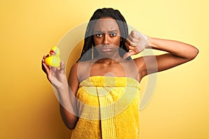 African american woman wearing shower towel holding toy duck over isolated yellow background with angry face, negative sign