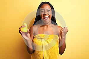 African american woman wearing shower towel holding toy duck over isolated yellow background screaming proud and celebrating