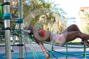 African American woman wearing sports clothes and white headphones, lying with her eyes closed listening to music in a playground