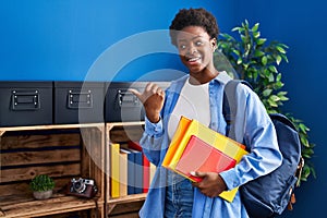 African american woman wearing student backpack and holding books pointing thumb up to the side smiling happy with open mouth