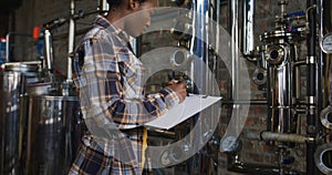 African american woman working at gin distillery checking equipment and writing on clipboard