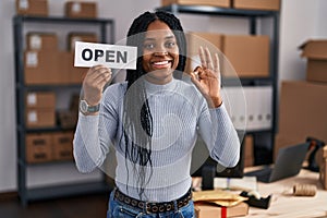 African american woman working at small business ecommerce holding open banner doing ok sign with fingers, smiling friendly