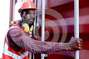 African American young engineer worker man wearing safety bright red vest and helmet, trying to open a shipping container door at