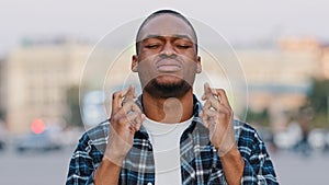 African American young hopeful man stands in city on street crossing fingers praying says prayer hopes for future