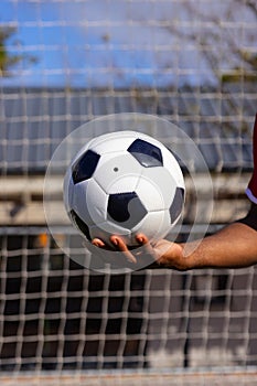 An African American young male athlete holding a soccer ball in front of a goal net on field outdoor