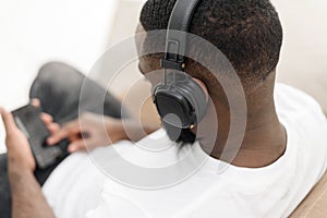 African american young male enjoying music in wireless headphones while sitting on sofa at home. Close up