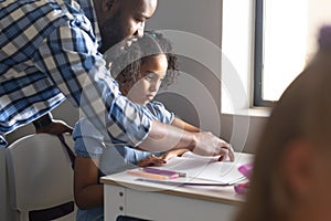 African american young male teacher teaching biracial elementary schoolgirl at desk in classroom