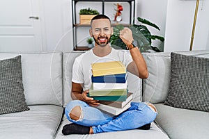 African american young man holding a pile of books sitting on the sofa angry and mad raising fist frustrated and furious while