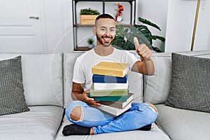 African american young man holding a pile of books sitting on the sofa approving doing positive gesture with hand, thumbs up
