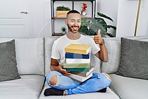African american young man holding a pile of books sitting on the sofa doing happy thumbs up gesture with hand