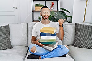 African american young man holding a pile of books sitting on the sofa pointing to the back behind with hand and thumbs up,