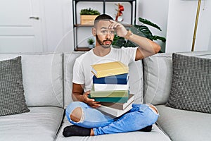 African american young man holding a pile of books sitting on the sofa worried and stressed about a problem with hand on forehead,