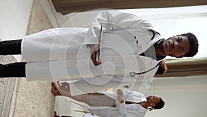 African american young man physician in white gown and stethoscope looking at camera and smiling in clinic. Women