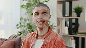 African american young man sitting on sofa, looking and smiling at camera in living room at home. Portrait of male