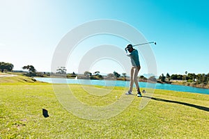 African american young man taking golf shot with club at golf course against lake and clear sky