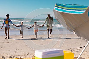 African american young parents holding son and daughter\'s hands while walking at beach against sky