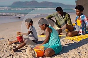 African american young parents sitting with son and daughter playing with pails and sand at beach