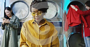 African American young pretty and stylish girl in yellow glasses standing in laundry service room and flipping pages of