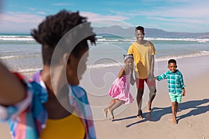 African american young woman looking at happy son, daughter and boyfriend at beach against sky