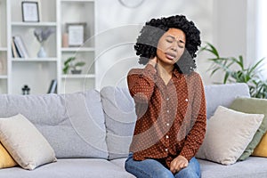 African American young woman sitting at home on sofa tired, closing eyes resting and doing neck massage, feeling pain